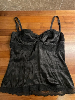 Dolce & Gabbana topp - Snygg svart linnetopp i satin med tunna axelband och spetsdetaljer vid byst och nederkant från Dolce & Gabbana. Toppen har en lätt glansig finish och ger en elegant look. Perfekt att styla med jeans eller kjol för en trendig outfit. Står L på lappen men skulle mer säga som en s/m. Sömmen har gått upp lite (se bild 3) går att fixa!