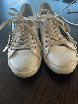Vita Charlotte Of Sweden - Säljer ett par vita sneakers i läder med snörning och dragkedja på sidan. Skorna har en klassisk låg profil och neutrala vita skosnören. Perfekta för en clean och stilren look.