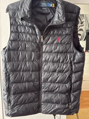 Svart dunväst från Polo Ralph Lauren - Snygg svart dunväst från Polo Ralph Lauren med quiltad design och dragkedja framtill. Västen har två sidofickor med dragkedja och en liten röd logga broderad på bröstet. Perfekt för lager-på-lager och streetstyle.