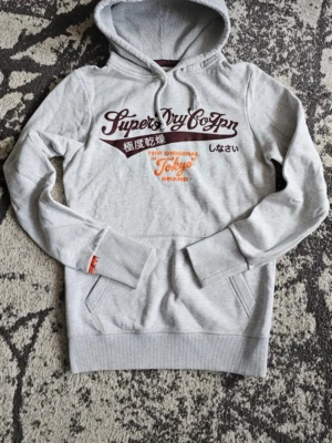 Grå hoodie Superdry Tokyo Brand - Snygg grå hoodie från Superdry med tryck i vinrött och orange på bröstet där det står 'Superdry Co Jpn' och 'Tokyo Brand'. Hoodien har huva med dragsko och en stor magficka. Perfekt för en avslappnad streetwear-look. Skriv om ni har frågor följ gärna detta konto för mer 