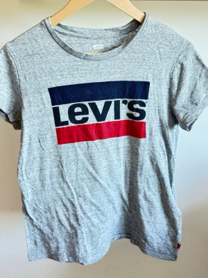 Grå Levi's t-shirt med logga strl XS - Snygg grå t-shirt från Levi's med klassisk stor logga i blått och rött framtill. T-shirten har rund halsringning och korta ärmar. Tillverkad i mjuk bomull för en bekväm känsla. Perfekt till jeans eller shorrts