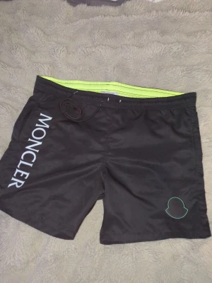 Svarta badbyxor från Moncler - (oanvända) snören kommer vid sidan finns inga på men de finns med )Snygga svarta badbyxor från Moncler med vit logotyp längs benet och en grön detalj på sidan. Insidan är fodrad med neongult meshfoder och midjan har dragsko för bästa passform. Perfekta för stranden eller poolen.