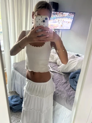 Vit långkjol med volangdetaljer - Säljer en superfin vit långkjol med flera volanglager och broderade detaljer. Kjolen har en luftig och flowy känsla, perfekt för varma dagar. Resår i midjan för skön passform och tillverkad i ett lätt material som känns bekvämt hela dagen.