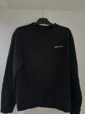 Svart sweatshirt från Axel Arigato - Säljer en svart sweatshirt från Axel Arigato med vit text 'ARIGATO ©2014' på bröstet. Tröjan har rund halsringning, ribbade muddar och är gjord i mjukt material. Perfekt för en clean och stilren look.