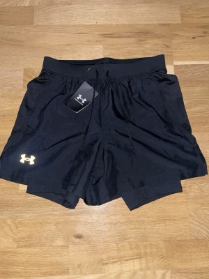 Svarta träningsshorts Under Armour - Snygga svarta träningsshorts från Under Armour med elastisk midja och dragsko. Shortsen har två sidofickor och ett vitt logotryck på vänster ben. Tillverkade i lätt och snabbtorkande material, perfekta för gym eller löpning.