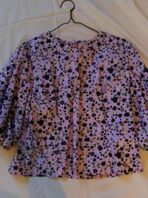 Lila blommig blus med puffärm - Söt blus i lila med svart och vit blommönster. Blusen har rund halsringning och puffiga långärmar som ger en trendig siluett. Tillverkad i ett lätt tyg som känns skönt mot huden. Perfekt för dig som vill ha en färgglad och unik look. Står stolek 44 men är mer som 40
