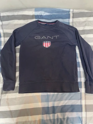 Mörkblå sweatshirt från GANT - Mörkblå sweatshirt från GANT med rund halsringning och lång ärm. Framtill finns GANT-logga och ett broderat sköldmärke i rött, vitt och blått. Tröjan har ribbade muddar vid ärmslut och nederkant. Perfekt för en avslappnad och stilren look.