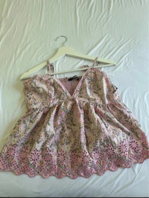 Blommig rosa linnetopp med spets - Supersöt rosa linnetopp med blommigt mönster och tunna axelband. Toppen har v-ringning, broderade spetsdetaljer längst ner och smock i ryggen för extra komfort. Perfekt för varma dagar och ger en romantisk vibe.