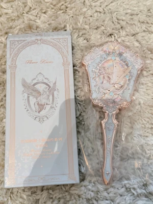 Flower knows Heaven Raven Hand Mirror - This is a decorative hand mirror with an ornate, vintage-inspired design. The mirror features a winged horse (Pegasus) motif in the center, surrounded by intricate gold and pastel embellishments. The packaging and the mirror both have elegant, classical artwork and text in both English and Chinese. The handle and frame are detailed with gold accents, giving it a luxurious and collectible appearance. This item is likely part of a themed collection, possibly inspired by fantasy or celestial motif