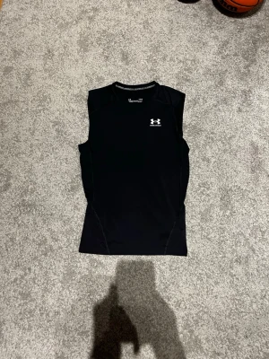 Svart ärmlös träningströja Under Armour - Snygg svart ärmlös träningströja från Under Armour med logga på bröstet. Tillverkad i ett stretchigt funktionsmaterial som andas, perfekt för intensiva träningspass eller gymmet. Tight passform och rund halsringning. Har använts 1-2 gånger.