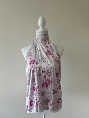 Ljusrosa halterneck blommig topp - Säljer en ljusrosa halterneck topp med blommigt mönster i rosa och lila toner. Toppen har hög hals med knyt i nacken och är ärmlös. Materialet känns glansigt och liknar satin