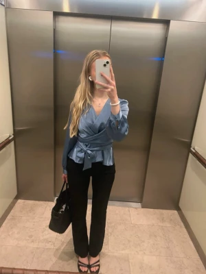Blå omlottblus med knytband - Snygg blå omlottblus med knytband i midjan och v-ringad hals. Blusen har långa ärmar och en lätt glansig finish som ger en elegant look. Perfekt att styla med svarta byxor eller jeans. Kan knytas både fram och bak.