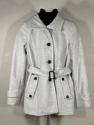 Burberry London Ljusgrå  ullkappa med bälte - Stilren ljusgrå ullkappa från Burberry London med knappar framtill och matchande bälte i midjan. Jackan har två stora fickor med lock och knappdetaljer samt klassisk krage. Materialet ser ut att vara en mjuk ullblandning som ger en elegant look. Perfekt för dig som vill ha en tidlös och snygg jacka.0109