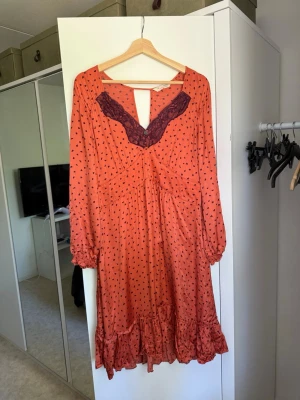 Orange långklänning med spetsdetalj - Snygg orange klänning med små svarta hjärtan, lila spets vid v-ringningen och volangkant nedtill. Klänningen har lång ärm och en feminin passform med markerad midja. Perfekt för dig som gillar romantiska detaljer och lekfulla mönster. Från märket Odd Molly i storlek 4 som enligt deras storlekguide motsvarar en storlek XL, men är liten i storlek. Kanske mer en L. Fin men tyvärr finns en massor av tråddragningar. 