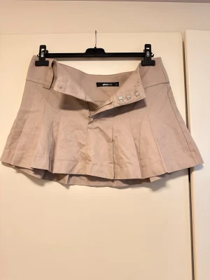 Beige kortkjol från Gina Tricot - Säljer en beige kortkjol från Gina Tricot med snygga silverfärgade knappar i midjan. Kjolen har en lätt utsvängd form och är gjord i ett mjukt bomullsmaterial. Perfekt att matcha med en crop top eller hoodie.