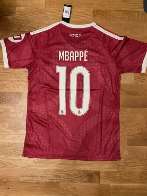 26/27 Vinröd Real Madrid Mbappé matchtröja - Snygg vinröd Real Madrid matchtröja med Mbappé och nummer 10 på ryggen. Tröjan har vita detaljer, klubbmärke och Adidas-logga på bröstet samt sponsortryck. Tillverkad i lätt och ventilerande material, perfekt för fotboll eller träning.