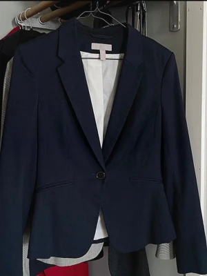 Mörkblå kavaj från H&M - Stilren mörkblå kavaj från H&M med figurnära passform och ett knappstängning framtill. Kavajen har smala slag, två fickor och är helfodrad med ljus insida. Perfekt för en snygg och uppklädd look. Notera viss kort längd.