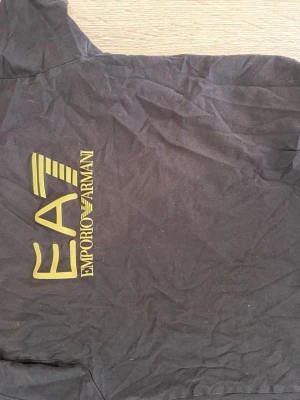 Svart T-shirt från EA7 Emporio Armani - Snygg svart t-shirt från EA7 Emporio Armani med gul logga på bröstet. Tillverkad i mjukt material som känns skönt mot huden. Perfekt för dig som vill ha en stilren och trendig look.