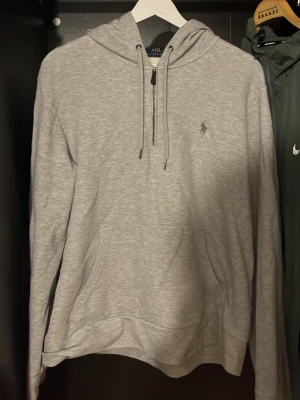 Ljusgrå hoodie från Polo Ralph Lauren - Ljusgrå hoodie med dragsko i huvan och klassisk Polo Ralph Lauren-logga broderad på bröstet. Tillverkad i mjukt bomullsmaterial och har en stor magficka framtill. Perfekt för en chill och avslappnad stil.