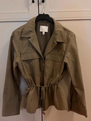 Grön jacka med snörning från H&M - Stilren jacka från H&M med markerad midja tack vare snörning. Jackan har stora bröstfickor, klassisk krage och långa ärmar. 