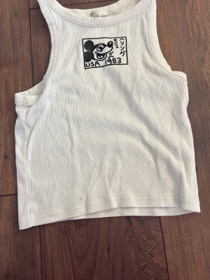 Vit ribbad linnetopp från H&M - Vit ribbad linnetopp från H&M med ett svart tryck framtill som föreställer en tecknad figur och texten 'USA 1983'. Toppen är ärmlös och har en avslappnad passform, perfekt för en cool och enkel stil.