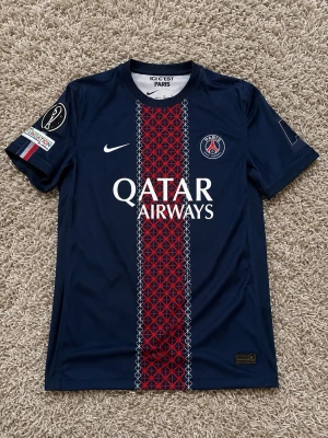 PSG matchtröja  - Säljer en blå PSG matchtröja från Nike med Qatar Airways-tryck på bröstet. Tröjan har ett rött och blått mönster i mitten, PSG-logga på vänster bröst och Nike-logga på höger. Korta ärmar och rund hals. Perfekt för fotbollsfans!