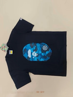 Svart BAPE t-shirt med blått camomönster - Säljer en svart t-shirt från BAPE med det ikoniska apansikte-trycket i blått camomönster på bröstet. T-shirten har korta ärmar och rund hals. Perfekt för dig som gillar streetwear och vill sticka ut med en unik design.