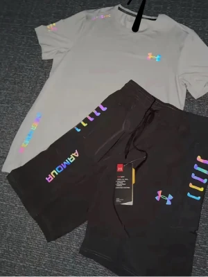 Under Armour shorts med regnbågsdetaljer - Säljer ett snyggt Under Armour shorts. plaggen har färgglada regnbågsdetaljer och logotyper. shortsen har dragsko i midjan. Perfekt för dig som gillar sportig och färgstark stil.