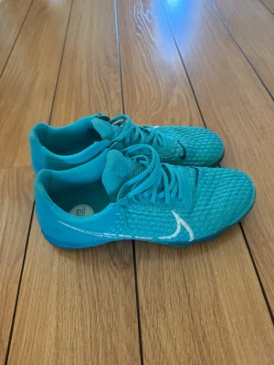 Turkosa fotbollsskor med vit detalj - Säljer ett par turkosa fotbollsskor med vit logga på sidan och svart detalj på tån. Skorna har snörning och ovandel i mesh-liknande material för bra ventilation. Yttersulan är platt och passar perfekt för spel på inomhusunderlag.