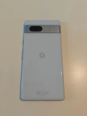 Google Pixel 7a  - Säljer en Google Pixel med stilren design i ljusblått glas och stor skärm. Telefonen har en modern look med en tydlig G-logga på baksidan och en svart kameramodul. Perfekt för dig som gillar Android och vill ha en snygg och snabb mobil. Skick syns på bilderna. Spricka framskärmen (knappt synlig)