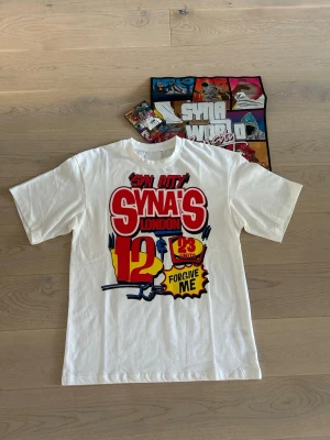 Synaworld t shirt - Vit t-shirt med stort, färgglatt tryck på framsidan med texten 'Syna's London' och grafiska detaljer i rött, gult och blått. Klassisk passform med rund hals och korta ärmar. Tillverkad i mjuk bomull, perfekt för en avslappnad streetwear-look.