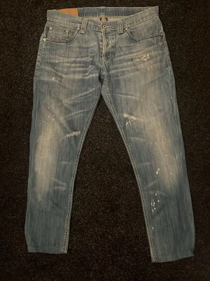 Ljusblå jeans från Dondup - Säljer ett par ljusblå jeans från Dondup med slitna detaljer och färgstänk för en cool, avslappnad look. Klassisk femficksmodell med raka ben och knappgylf. Tillverkade i denim med snygg tvättad finish.