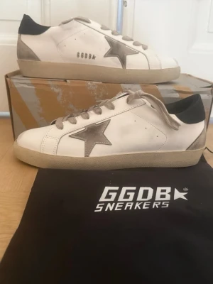 Vita GGDB sneakers med grå stjärna - Säljer ett par vita GGDB sneakers med grå stjärna på sidan och beige detaljer. Skorna har svarta hälpartier och beige snören. Klassisk låg modell i läder med slitna detaljer för en cool look. Perfekta för dig som gillar streetstyle och unika sneakers.