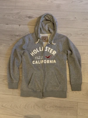 Grå Hollister hoodie - Grå hoodie från Hollister med vit broderad text 'Hollister 1922 California' och röd logga på bröstet. Tröjan har dragkedja framtill, två fickor och en huva med vita snören. Tillverkad i mjukt sweatshirtmaterial, perfekt för en avslappnad stil. Storlek: Står XL men passar mer som M. 
