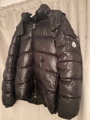 Svart dunjacka från Moncler - Snygg svart dunjacka från Moncler med glansig finish, hög krage och avtagbar huva. Jackan har dragkedja framtill, fickor med dragkedja och en ficka med lock på ärmen där Moncler-loggan sitter. Perfekt för kalla vinterdagar. Jackan är i storlek M (och är ganska stor på mig som är 164cm)      Orginalpåse och prislappar kommer med!  Pris kan diskuteras  Orginalpris / köpt för 11.000kr