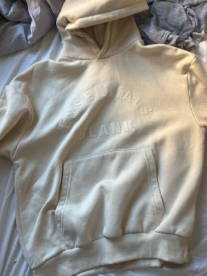 Beige hoodie Essentials Blank - Snygg beige hoodie med huva och stor magficka framtill. På bröstet står det 'ESSENTIALS BLANK' i upphöjd text. Hoodien har ribbade muddar vid ärmslut och nederkant. Perfekt för en avslappnad och trendig look.