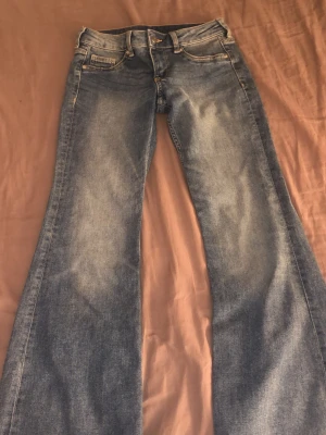 Blå bootcut jeans med slitningar - Säljer ett par blå bootcut jeans med snygga slitningar framtill. Jeansen har klassisk femficksdesign, normal midja och utsvängda ben. Perfekta för dig som gillar retrostil och vill ha ett par jeans som sticker ut lite extra.