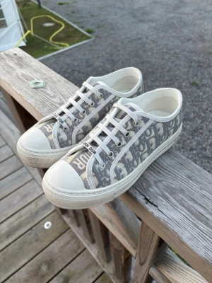 Gråvita Dior sneakers med monogram - Säljer ett par gråvita sneakers från Christian Dior med klassiskt Dior-monogram i tyg. Skorna har vita snören, gummisula och tåhätta samt detaljer med logotyp på plösen. Kartong samt påse medföljer. Endast använda ungefär 4 gånger då dom är lite för stora för mig. Jag har köpt dom här på Plick så jag är osäker på om dom är äkta eller inte men dom ser ut som dom ska och qr kod finns.