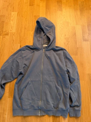 Blå zip hoodie från Weekday - Säljer en blå hoodie från Weekday i storlek M. Hoodien har dragkedja framtill, två stora fickor och en klassisk huva med snören. Tillverkad i mjukt bomullsmaterial som är skönt mot huden. Perfekt för en avslappnad och chill stil.