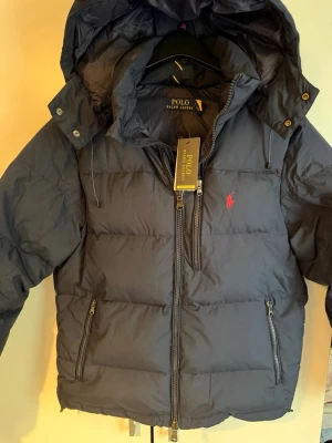 Polo Ralph Lauren Vinterjacka - Snygg mörkblå dunjacka från Polo Ralph Lauren med huva, dragkedja framtill och tre praktiska fickor med dragkedja. Jackan har det klassiska röda Polo-logotypen på bröstet och en puffig, varm design perfekt för kalla vinterdagar.