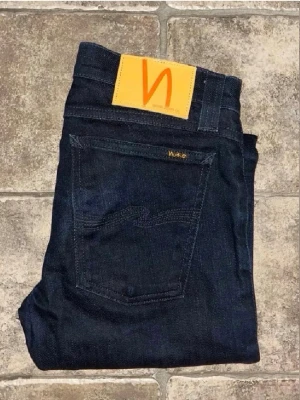 Nudie jeans  - Snygga mörkblå jeans från Nudie i modellen Tight Long John. De har en slim passform med smala ben och klassisk femficksdesign. Materialet är 99% bomull och 1% elastan för lite stretch. Midja 35cm, längd 105cm.  Ifall det inte finns någon bild i annonsen där jag bär plaggen jag säljer så är det för att jag ej kan ha storleken heller passformen, så kolla måtten för storlek och googla på modellnamnet för passform 😉 (n3)