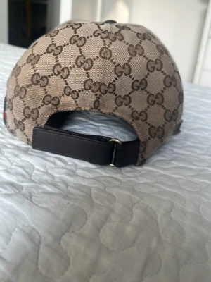 Beige keps med GG-mönster från Gucci - Säljer en beige keps från Gucci med det klassiska GG-monogrammet i brunt över hela kepsen. Kepsen har justerbart band bak i svart läder och gröna/röda ränder på sidan. Materialet är canvas och kepsen har böjd skärm.