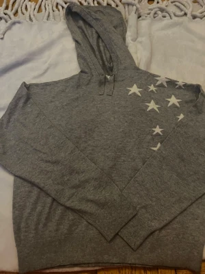 Grå stickadhoodie med vita stjärnor - Säljer en grå stickad hoodie med vita stjärnor över ena axeln och bröstet. Hoodien har huva med dragsko och långa ärmar. Tillverkad i mjukt  stickat material som passar perfekt för en avslappnad stil. Väldigt fin nästan aldrig använd. 