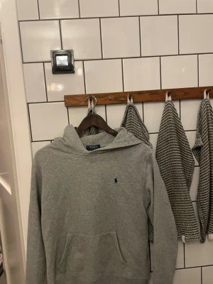 Grå hoodie från Polo Ralph Lauren - Snygg grå hoodie från Polo Ralph Lauren med klassisk huva och känguruficka framtill. Diskret blå logga broderad på bröstet. Tillverkad i mjukt bomullsmaterial som är perfekt för chill dagar. Enkel och stilren design som funkar till det mesta.