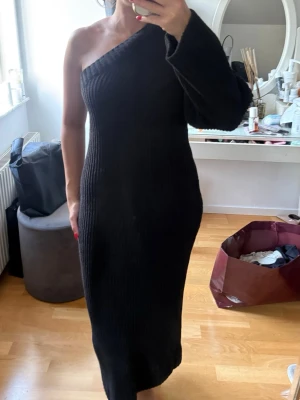 Svart oneshoulder stickad långklänning - Säljer en svart ribbad långklänning med oneshoulder-design och lång ärm på ena sidan. Klänningen är stickad och har en tight passform som framhäver figuren. Perfekt för dig som vill ha en stilren och trendig look.