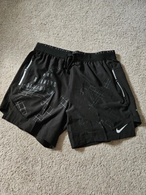 Svarta löparshorts från Nike - Snygga svarta löparshorts från Nike med reflekterande detaljer och tryck. Shortsen har dragkedjefickor på sidorna, elastisk midja med snörning och Nike-logga framtill. Perfekta för träning och sportiga outfits.