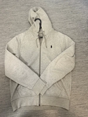  Ralph Lauren hoodie - Säljer en stilren ljusgrå hoodie med dragkedja och huva. Hoodien har fickor framtill, ribbade muddar och en liten broderad logga på bröstet. Perfekt för en avslappnad look och enkel att matcha med det mesta.