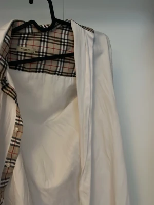 Vit skjorta med rutiga detaljer Burberry - Klassisk vit skjorta från Burberry med ikoniska rutiga detaljer på insidan av kragen och knappslån. Skjortan har lång ärm, knappar framtill och är tillverkad i mjuk bomull. Perfekt för en stilren och tidlös look.
