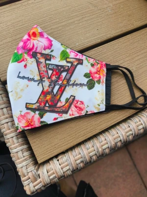 Blommigt munskydd med LV-tryck - Snyggt munskydd i vitt tyg med färgglada blommor och stort LV-tryck framtill. Svarta elastiska band för öronen. Perfekt accessoar för dig som vill sticka ut och addera lite lyxig känsla till din look.knappt använd och tvättad 