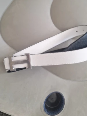 Vitt skärp med silverfärgat spänne - Snyggt vitt skärp i skinn med ett stilrent, silverfärgat H-format spänne. Perfekt accessoar för att lyfta din outfit och ge en clean look. Bältet har en slät yta och klassisk design.